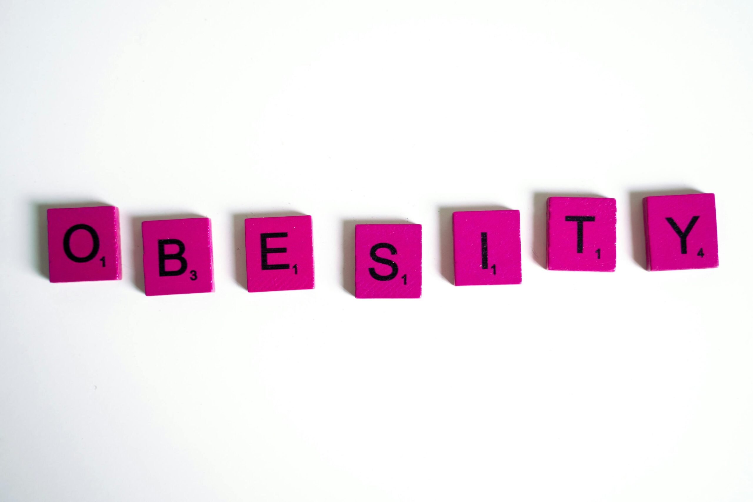 Purple Scrabble tiles spelling 'obesity' on a white surface, conceptual still life.